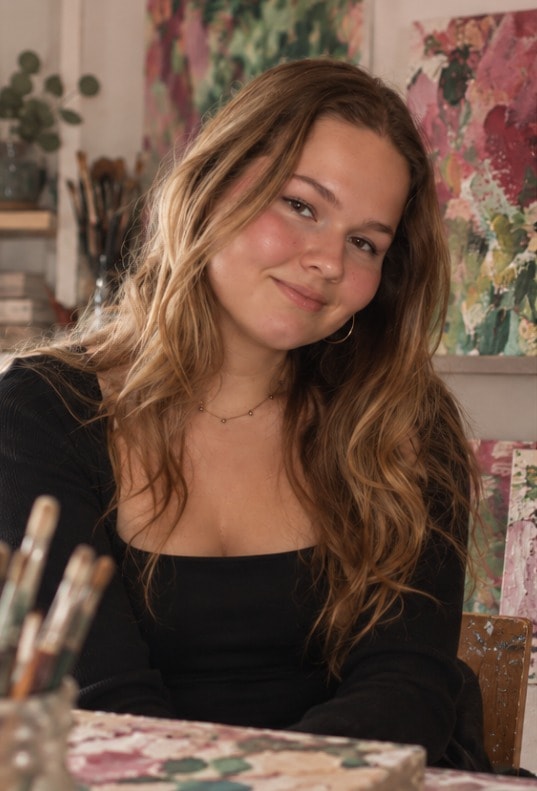 Daphne Merel, lachend, aan een tafel in haar atelier, met kwasten in een pot en kleurrijke schilderijen achter haar.