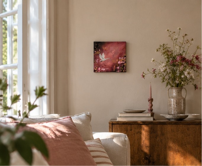 Flying Swan boven een houten dressoir in een zonnige woonkamer, naast een vaas met veldbloemen, een roze kandelaar en een bank met roze kussens.