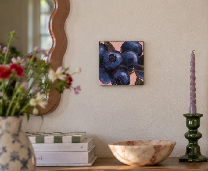 Blueberries aan een lichte muur boven een dressoir met een vaas veldbloemen, een gemarmerde schaal en een lila kaars in een groene kandelaar, naast een golvende spiegel.