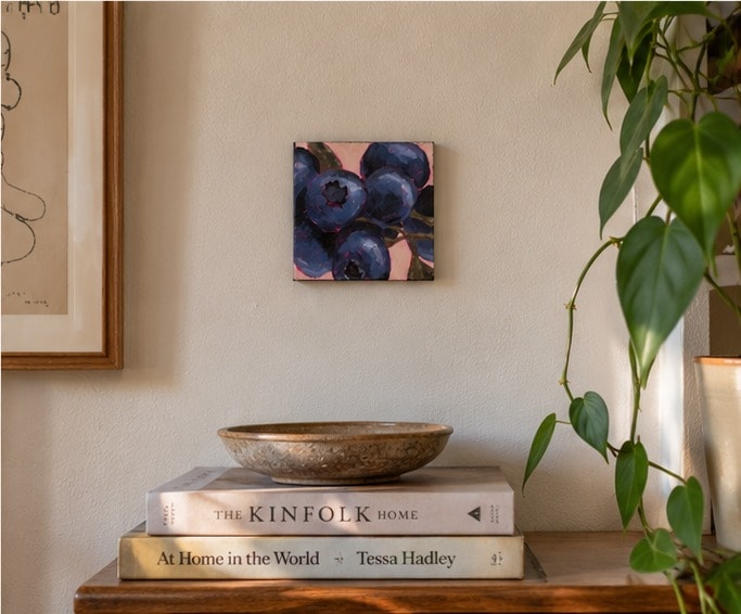 Blueberries aan een lichte muur boven een stapel boeken met een stenen schaal, naast een ingelijste lijntekening en een hangende plant.