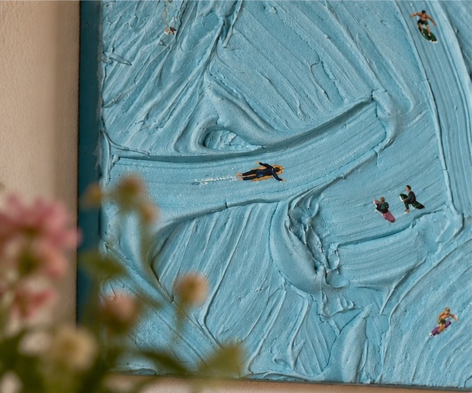 Close-up van Catching Waves schuin van opzij: kleine zwemmers en surfers op ruggen van dikke turquoise verf, met onscherpe roze bloemen ervoor.